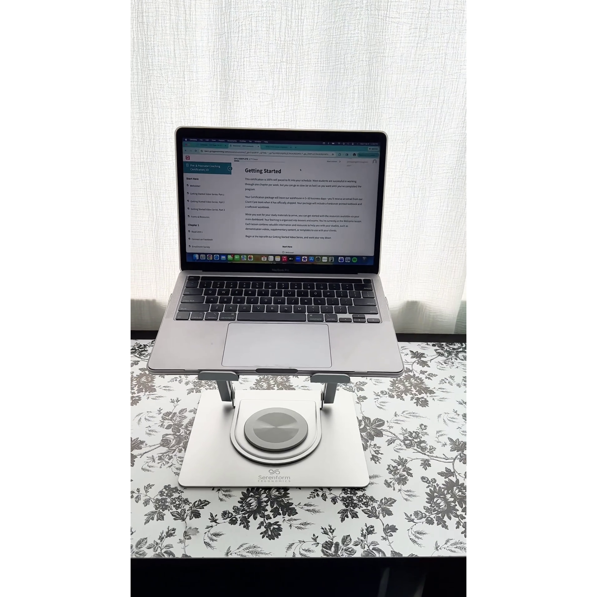 Front view of laptop placed on Serenform Elevate Laptop Stand on a floral-patterned tablecloth with a white curtain background